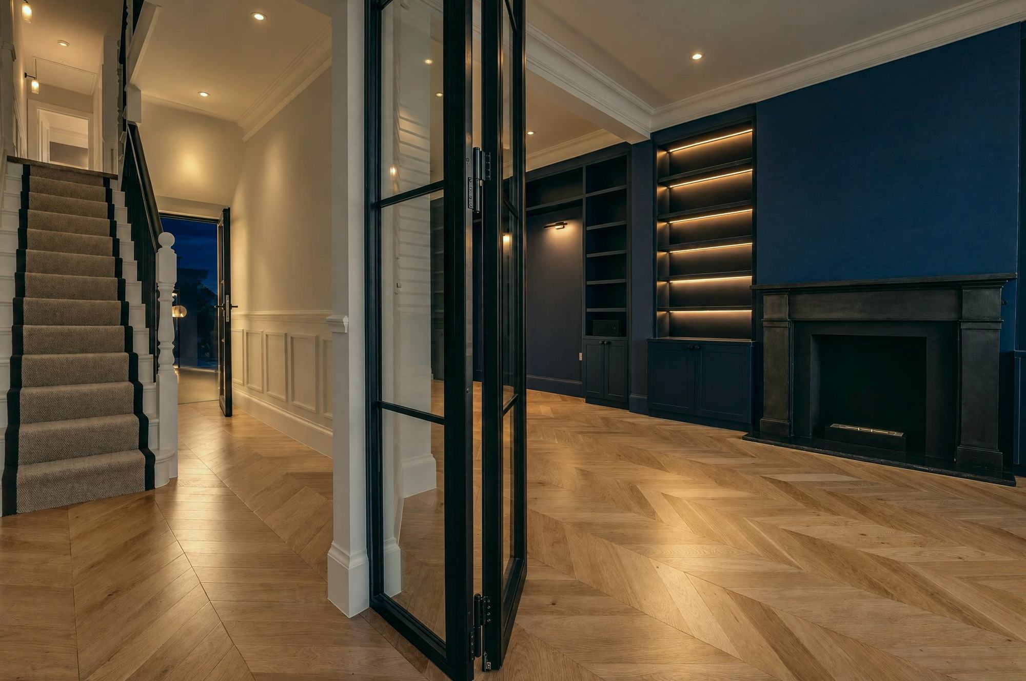Black steel folding glazed doors between the hall and a navy library in the evening